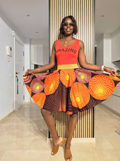 A woman wearing a vibrant Horizon Ankara Skirt with an orange and yellow pattern, styled casually in a modern setting.